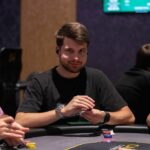 Ein Poker-Tisch mit grünem Filz, umgeben von Spielern, die Karten halten und Pokerchips stapeln. In der Mitte des Tisches liegt eine Gemeinschaftskarte. Die Spieler sitzen konzentri