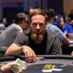 Ein Mann spielt Poker an einem Tisch mit grüner Filzdecke und Chips. Er hält Karten in der Hand und betrachtet sie konzentriert, während andere Spieler im Hintergrund zu sehen sind.