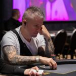 Ein Mann sitzt an einem Pokertisch und hält seine Karten mit ernstem Gesichtsausdruck. Ein Stapel Chips liegt vor ihm, während andere Spieler im Hintergrund zu sehen sind.