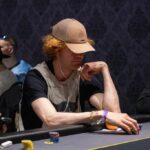 Ein Mann und eine Frau sitzen an einem Pokertisch und spielen Poker. Der Mann hat eine Handkarte in der Hand und schaut konzentriert auf seine Karten. Die Frau schaut ihn mit einem Lächeln an und
