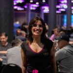 Ein Mann in einem schwarzen Anzug und einer roten Krawatte sitzt an einem Pokertisch und hält seine Spielkarten in der Hand. Im Hintergrund sind Chips und weitere Spieler zu sehen.