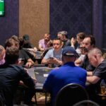 Ein Mann und eine Frau sitzen an einem Pokertisch und spielen eine Partie Poker. Der Mann hält Karten in der Hand und schaut konzentriert auf den Tisch, während die Frau mit ernstem Gesichtsaus