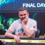 Ein Mann in einem schwarzen Anzug sitzt an einem Pokertisch und hält seine Karten. Andere Spieler sind im Hintergrund zu sehen.