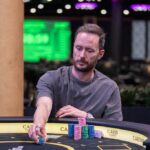 Ein Mann spielt Poker an einem Tisch in einem eleganten Casino. Er hat eine ernste Miene und hält Karten in der Hand, während er von anderen Personen umgeben ist. Die Atmosphäre ist gespannt und glamour