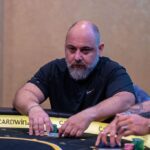 Ein Mann spielt Poker an einem eleganten Tisch mit grüner Filzdecke und hält dabei eine Hand mit Spielkarten. Im Hintergrund sind weitere Spieler zu sehen, die konzentriert ihre Karten betrachten