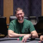 Ein Mann sitzt an einem Pokertisch und hält zwei Spielkarten in der Hand. Er trägt einen Anzug und eine dunkle Sonnenbrille. Im Hintergrund sind Pokerchips und andere Spieler zu sehen.