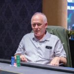 Ein Mann und eine Frau sitzen an einem Pokertisch und spielen Karten. Der Mann hat eine Hand mit Karten in der Hand, während die Frau mit ernstem Gesichtsausdruck ihre Karten betrachtet. Der T