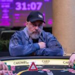 Ein Mann spielt Poker an einem Tisch mit grünen Filz und Chips. Er hat eine Hand aus Karten in der Hand und betrachtet konzentriert seine Gegner.