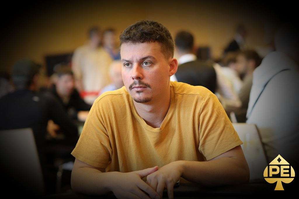 Ein Mann in einem schwarzen Anzug sitzt an einem Pokertisch und zeigt triumphierend seine Karten. Im Hintergrund sind andere Pokerspieler und ein Dealer zu sehen.