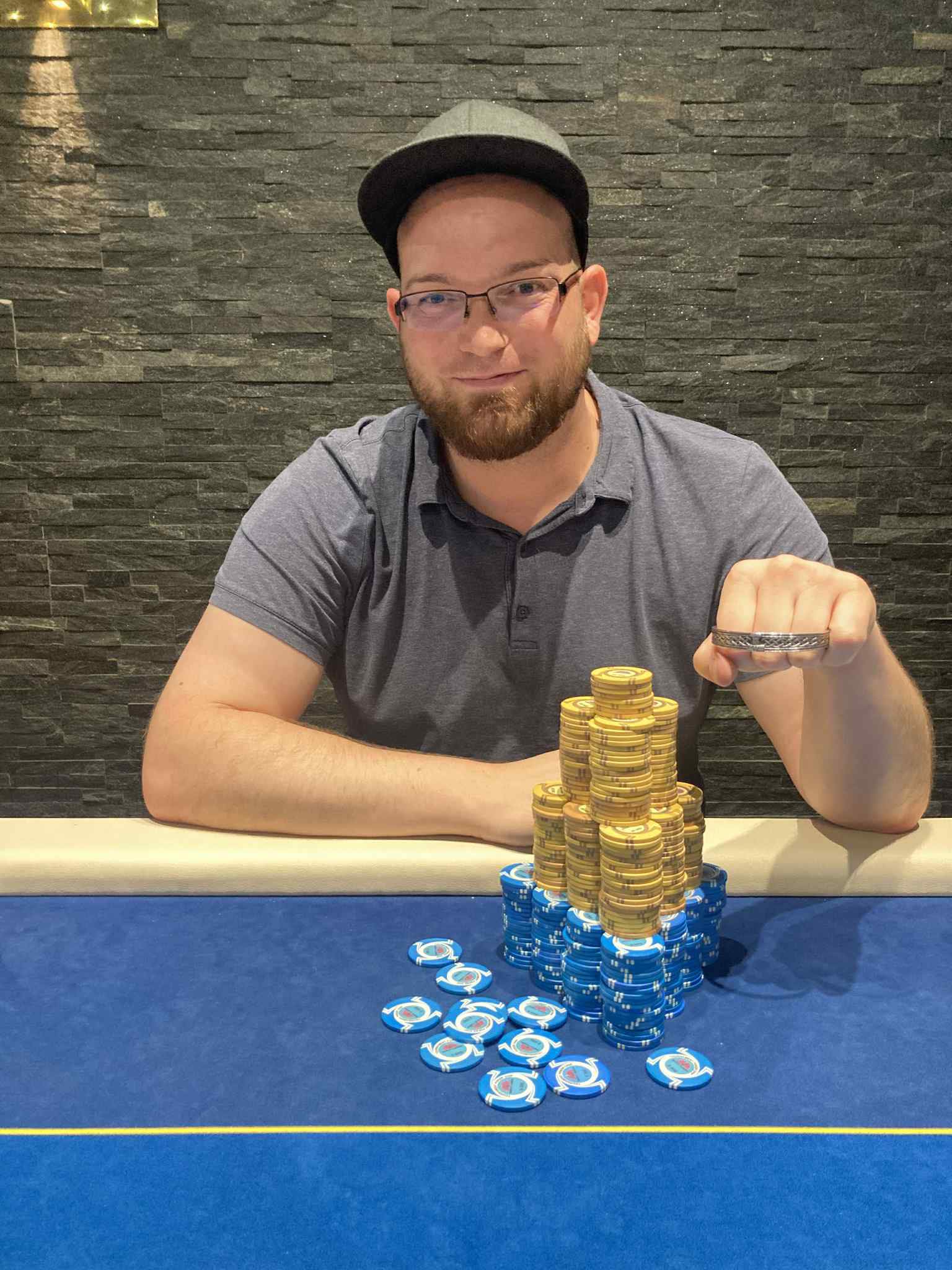 Ein Mann sitzt an einem Pokertisch und schaut konzentriert auf seine Karten. Er hat dunkles Haar und trägt ein schwarzes Hemd. Im Hintergrund sind andere Spieler und Spielchips zu sehen.