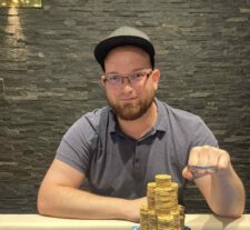 Ein Mann sitzt an einem Pokertisch und betrachtet konzentriert seine Karten. Im Hintergrund sind andere Spieler und Chips zu sehen.