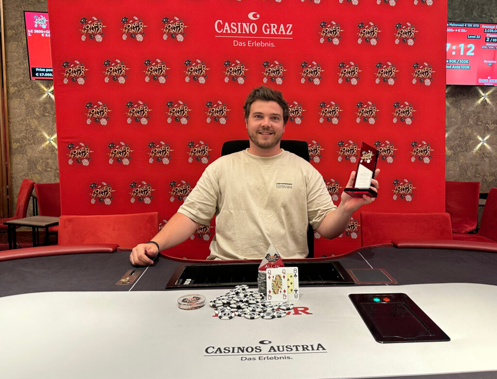 Ein Mann mit einem breiten Grinsen hält stolz einen Pokal in die Luft, während er von anderen Pokerspielern umgeben ist.