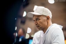 Ein Mann in einem schwarzen Anzug und einer schwarzen Mütze sitzt an einem Pokertisch und schaut konzentriert auf seine Karten. Im Hintergrund sind andere Spieler und Zuschauer zu sehen.