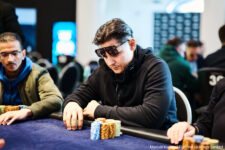 Ein Mann sitzt an einem Pokertisch und betrachtet nachdenklich seine Karten. Im Hintergrund sind andere Spieler und Pokerchips zu sehen.