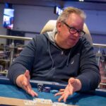 Ein Mann sitzt an einem Pokertisch im Casino. Seine Karten hält er verdeckt in der Hand, während er konzentriert auf den Tisch schaut. Im Hintergrund sind weitere Spieler und Dealer zu sehen