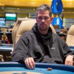 Ein Mann sitzt an einem Pokertisch während eines Turniers im Casino. Er hat eine Sonnenbrille auf und stützt seinen Kopf mit der Hand. Im Hintergrund sind andere Spieler und Dealer zu sehen.