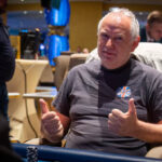 Ein Gruppenfoto von Pokerspielern, die an einem Turnier teilnehmen, während sie an einem Tisch sitzen und Karten spielen. Es sind Männer und Frauen verschiedener Altersgruppen zu sehen, die konzentriert