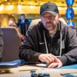 Ein Mann sitzt an einem Pokertisch während eines Turniers im "The Festival Rozvadov Mixed Games Main Event". Er hat eine Sonnenbrille auf und konzentriert sich auf seine Spielkarten, umgeben von anderen