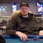 Ein Mann in einem dunkelblauen Hemd sitzt an einem Pokertisch und betrachtet seine Karten. Im Hintergrund sind weitere Pokerspieler und Dealer zu sehen. Das Bild wurde bei "The Festival Rozvad