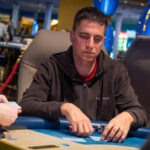 Ein Mann sitzt an einem Pokertisch während eines Turniers. Er hält Karten in der Hand und konzentriert sich auf das Spiel. Im Hintergrund sind andere Spieler und Dealer zu sehen.