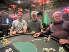 Ein glücklicher Mann in einem schwarzen Anzug und mit Sonnenbrille spielt Poker und hält zwei Karten in der Hand, während er lächelt. Im Hintergrund sind Pokerchips und ein grüner Fil