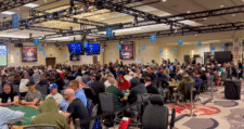 Ein Bild von einer Menschenmenge, die an einem Tisch sitzt und Poker spielt. Es herrscht eine aufgeregte Atmosphäre in einem großen Veranstaltungsraum während der World Series of Poker (WSOP)