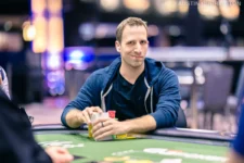Ein Mann in einem eleganten Anzug sitzt an einem Pokertisch und hält seine Karten mit einem selbstbewussten Blick. Um ihn herum sind andere Spieler zu sehen, die gespannt auf ihre Karten schauen.