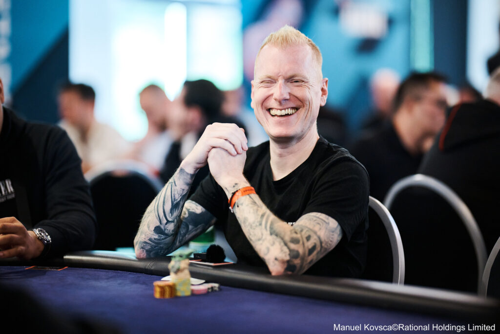 Ein männlicher Pokerspieler sitzt an einem Tisch während eines Turniers in Namur. Er konzentriert sich auf seine Karten und um ihn herum befinden sich andere Spieler.