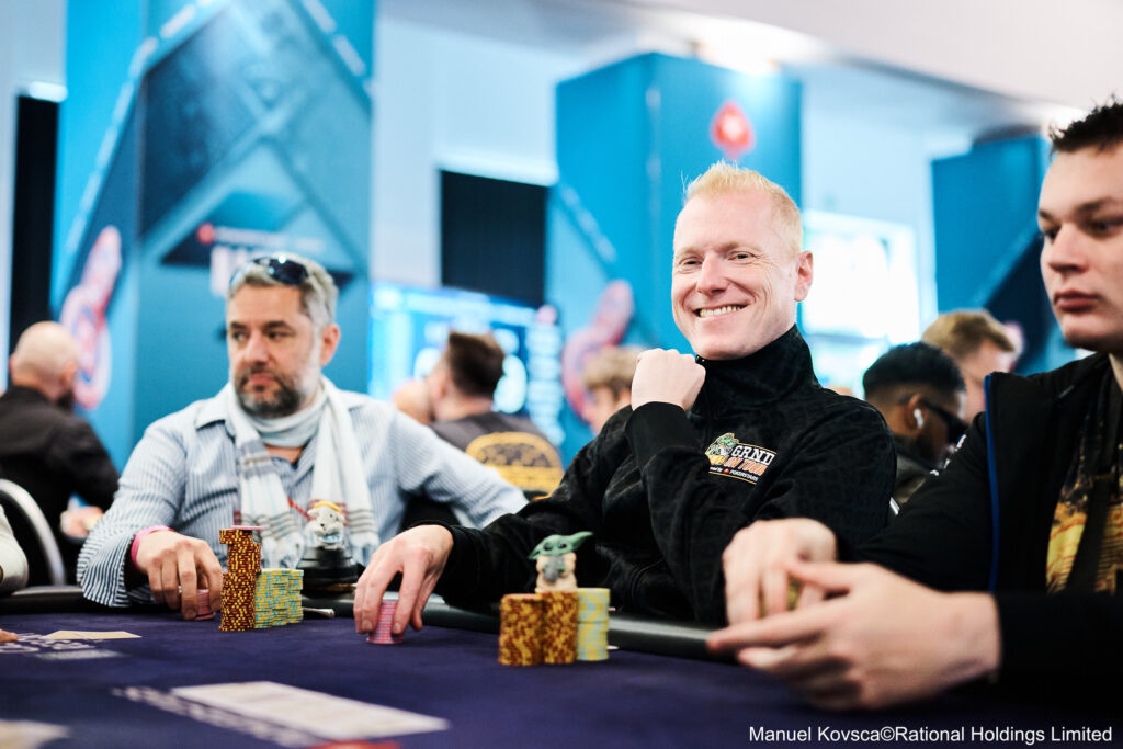 Ein Mann in einem schwarzen Anzug und einer schwarzen Mütze sitzt an einem Pokertisch und hält seine Karten in der Hand. Im Hintergrund sind andere Spieler und Spieltische zu sehen.