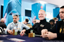 Ein Mann in einem schwarzen Anzug und einer schwarzen Mütze sitzt an einem Pokertisch und hält seine Karten in der Hand. Im Hintergrund sind andere Spieler und Spieltische zu sehen.